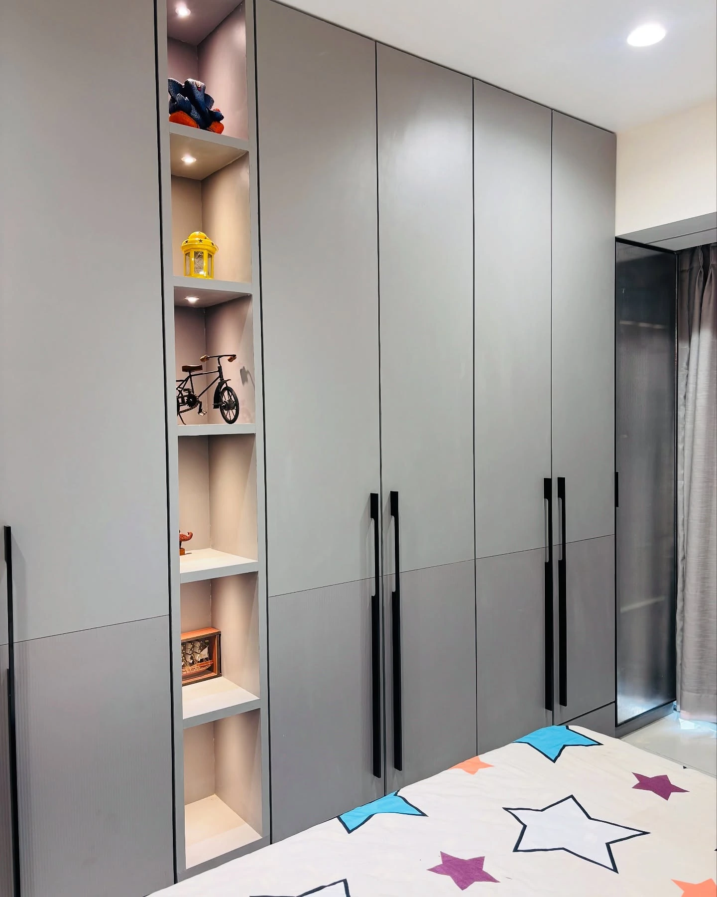 Matte grey kids wardrobe with open display niche shelves, black vertical handles, and recessed lighting in a modern children's bedroom