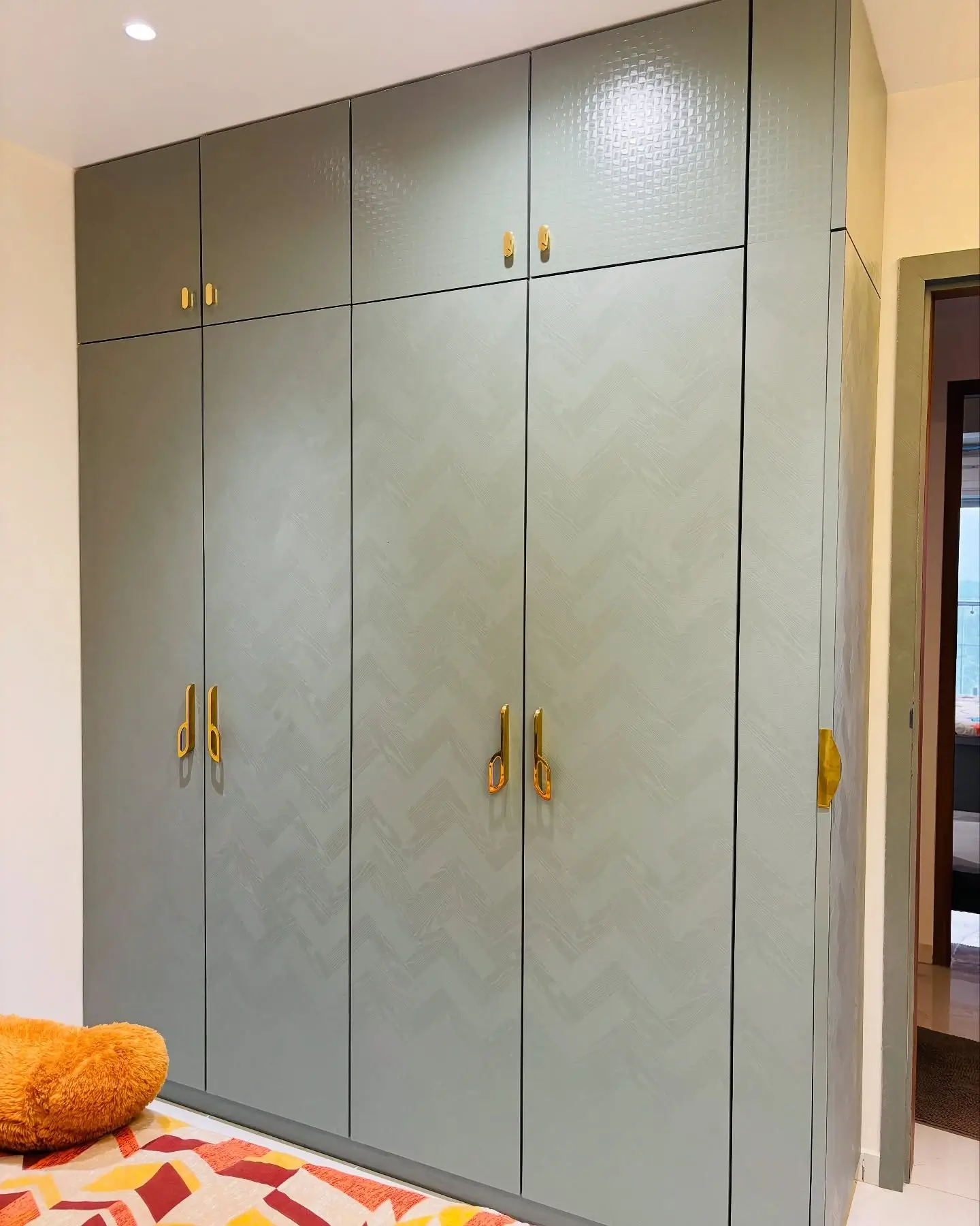 Sage green full-height wardrobe with chevron textured shutters, gold designer handles, and basketweave finish loft cabinets in a modern bedroom