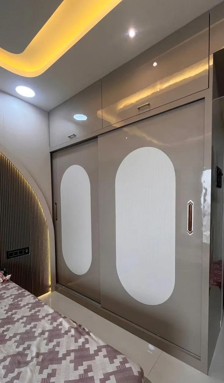 Modern Sliding Wardrobe with Glossy Finish and LED Lighting This sleek and modern sliding wardrobe design combines glossy beige panels with elegant white accents to create a luxurious, minimalistic look. The wardrobe features smooth, curved sliding doors that are highlighted with LED lighting, adding a touch of sophistication and warmth to the room. The clean design is perfect for contemporary bedrooms, providing ample storage space without compromising on style. Wardrobe Design Details: Type: Sliding Door Wardrobe with Glossy Panels and LED Lighting Doors: 2 Sliding Doors with Beige Glossy Panels and White Curved Accents Wardrobe Dimensions (WxDxH): Approx. 7 x 2.5 x 8 feet Style: Modern Minimalistic with Glossy Finish and Soft Curved Design Colour: Beige (Main Panels), White (Accents), Soft Yellow (LED Lighting) Wardrobe Shutters: High-gloss laminate finish with soft curved accents Special Features: Glossy beige finish enhances the modern, sleek look while making the space appear larger. Curved white accents on the sliding doors add a soft, contemporary design element. Integrated LED lighting under the wardrobe creates a warm glow, enhancing the visual appeal. The sliding door mechanism ensures smooth movement while saving space in smaller rooms. Ideal for modern bedrooms, combining both style and storage efficiency. Design Benefits: The combination of beige gloss and white accents adds an elegant and calming atmosphere to the room. LED backlighting under the wardrobe creates a cozy and inviting ambiance. Sliding doors save valuable space while offering easy access to the wardrobe’s contents. The clean, minimalist design seamlessly integrates with contemporary interiors, adding a stylish touch without clutter. Perfect for modern bedrooms or living areas, this wardrobe design maximizes storage and visual appeal.