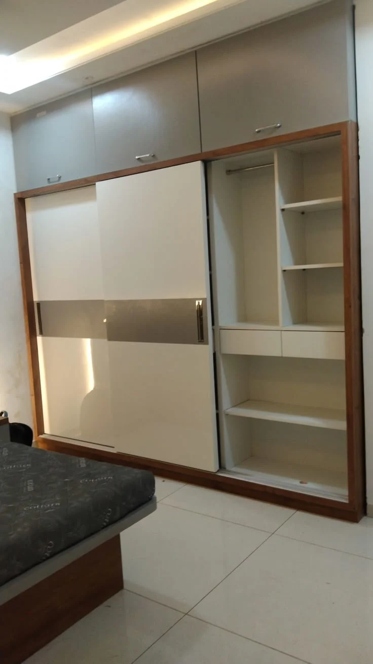 Grey and White Sliding Wardrobe with Wooden Accents Design Details: Unit Size (WxDxH): Customizable Style: Contemporary Minimalist with Elegant Accents Colour: Grey and White with Wooden Tones and Silver Inserts Finish: Glossy Laminate with Sleek, Clean Lines Storage Features: This modern grey and white sliding wardrobe is the perfect blend of contemporary style and practicality, ideal for Noida apartments. The wardrobe features a stylish grey and white sliding door with a silver insert, adding a sleek and sophisticated touch. The wooden frame complements the design, infusing warmth into the otherwise minimalist look. The upper cabinets offer spacious storage for bedding or accessories, while the inner shelves and drawers provide ample space for organizing clothes and other essentials. Special Features: The contrast of grey and white tones with wooden accents creates a chic and timeless design. The sliding doors maximize space efficiency, making it perfect for compact rooms in Noida sectors like Sector 34 and Sector 72. Well-designed compartments, including upper cabinets and lower shelves, make organizing easy and accessible. The glossy finish and silver insert add a luxurious, modern touch, making it suitable for urban Noida homes. This wardrobe design is a perfect choice for modern homes in Noida, offering both style and storage in a functional, space-saving design.